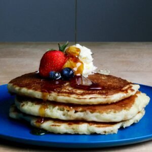 Stack of fluffy pancakes topped with syrup, strawberries, blueberries, and cream on a blue plate.
